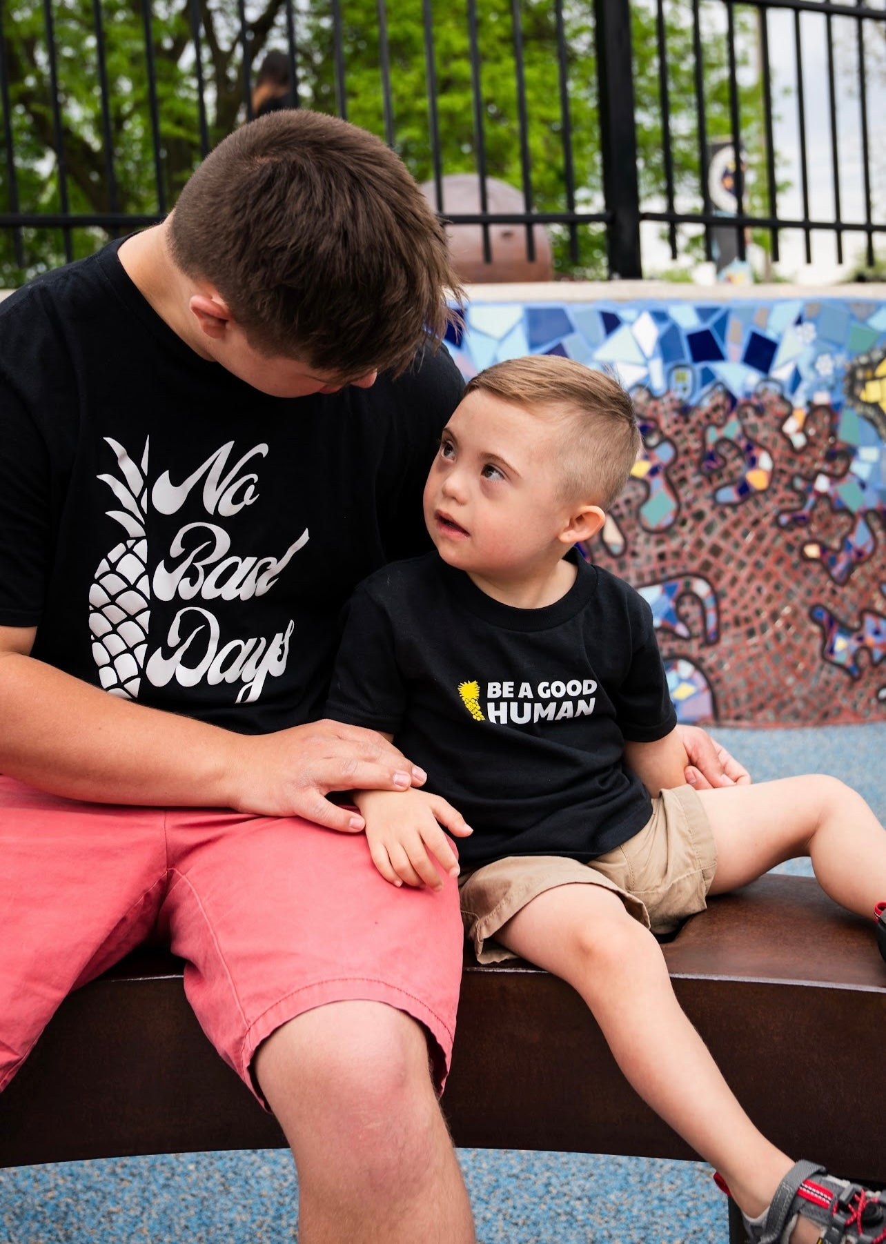 Nate Simon and child sitting together wearing black t-shirts with text, outdoor setting.